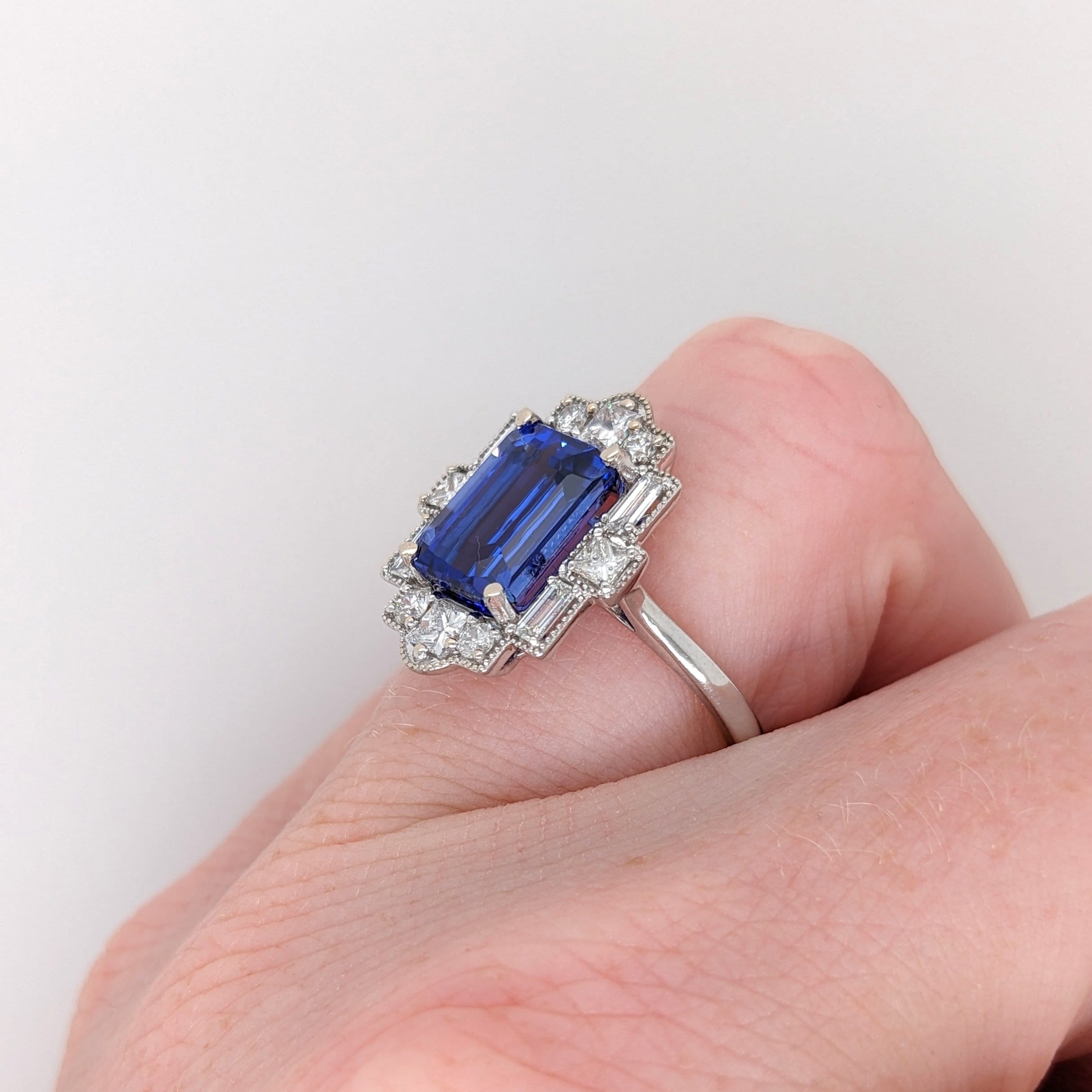emerald cut tanzanite 9x7mm blue natural gemstone and diamond ring in 14k white gold with milgrain detail, art deco style with a straight shank and halo of 4 baguette diamond accents 4 princess diamond accents and 4 round diamond accents